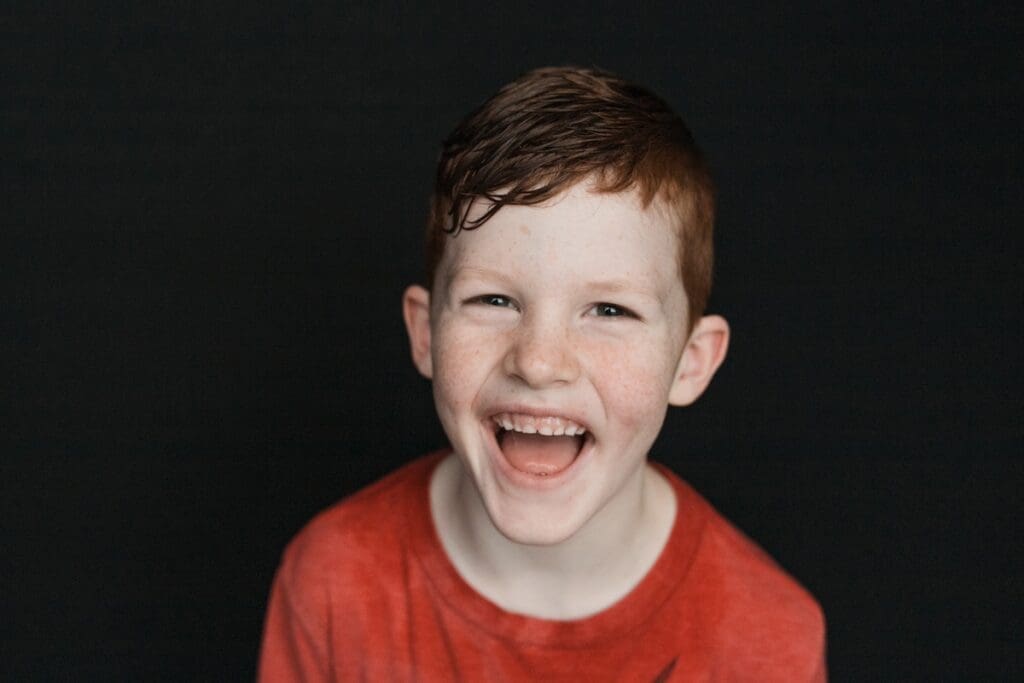 Boy giggling in front of a dark background showing pure joy and wearing a red t-shirt.