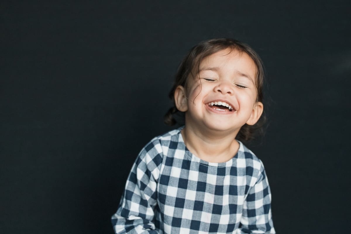 Young girl wearing a navy blue gingham check shirt laughs joyfully against dark background.