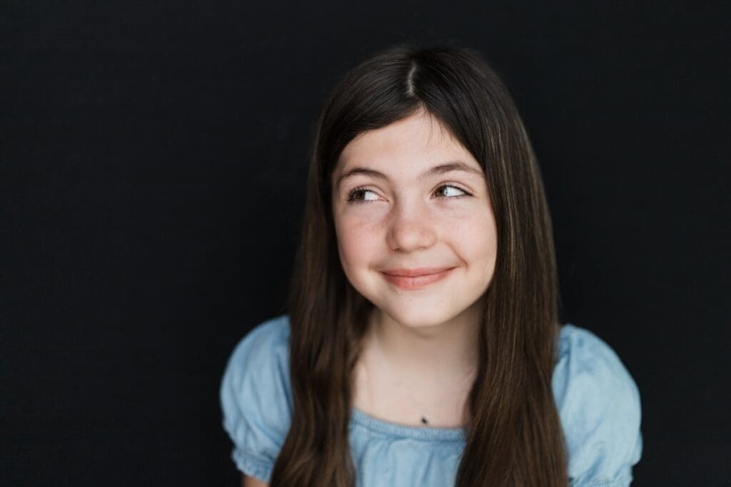A tween girl in a light blue top smiles while glancing upward against a dark background.