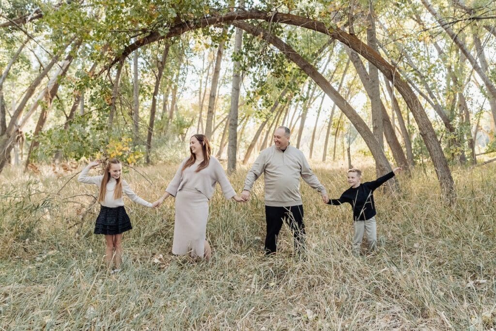 A family holds hands and walks together through a a field with tall trees for a family photo session.