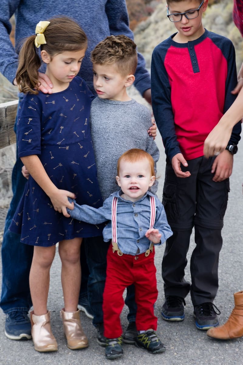 Children in coordinated casual outfits stand together outdoors, with a toddler in red suspenders at the center.