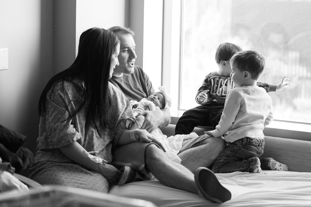 A candid black and white photograph shows a family spending time together with their new baby on a bed next to a sunlit window.