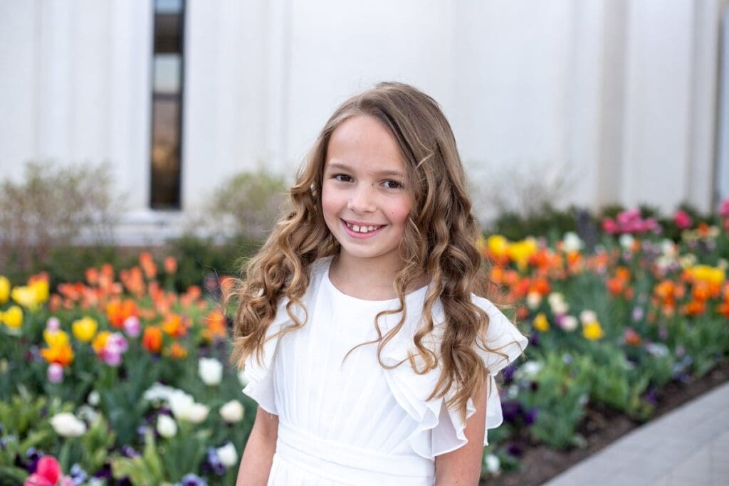 A young child in a white baptism dress smiles near a colorful tulip garden with orange and pink flowers blooming during her photo session with flowers