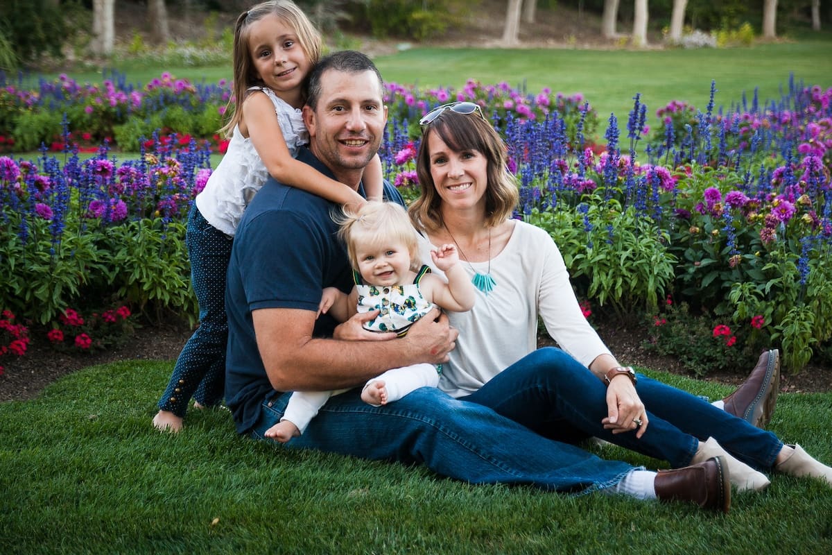 A young family sits together during a Utah photo session on grass surrounded by vibrant purple and pink flowers in a garden setting.
