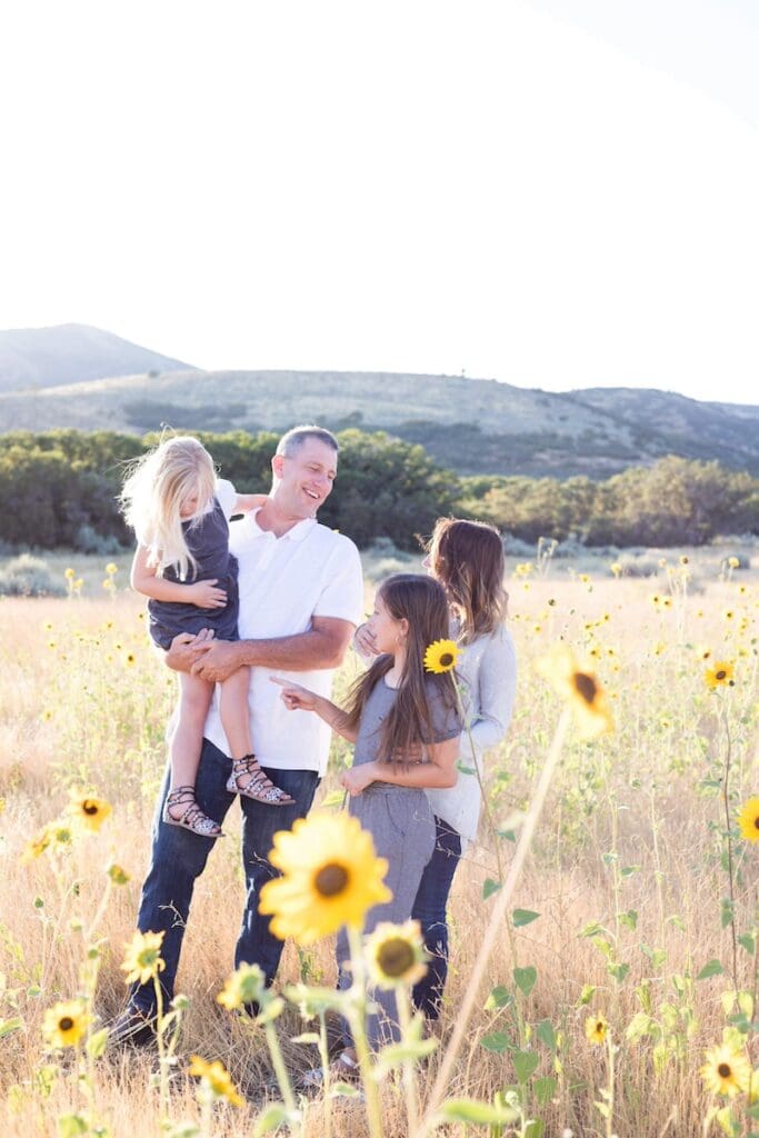 A family enjoys quality time together in a sunlit field of sunflowers with mountains in the background.