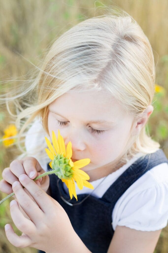 A gentle moment during a Utah photo session with flowers as a girl in a navy dress smells a bright yellow sunflower.