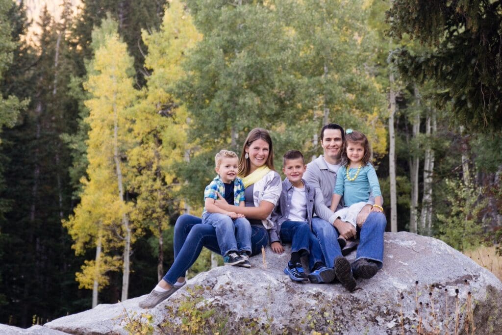 A family sits together on a large boulder in Big Cottonwood Canyon wearing teal, gray, and little pops of yellow.